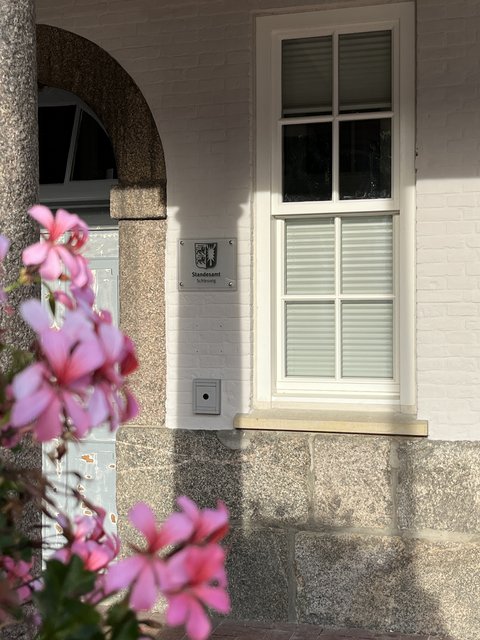 Auf dem Bild ist der Eingang des Standesamtes Schleswig mit dem Schild "Standesamt Schleswig" samt Wappen zu sehen. Im Vordergrund im Anschnitt ist eine Säule und rosa Blüten.